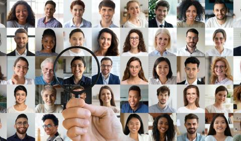 Une main passe une loupe sur un trombinoscope d'entreprise illustrant la diversité de genre, d'origine géographique, d'âge, etc.