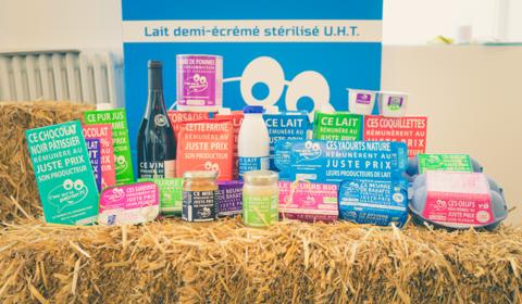 Différents produits alimentaires - produits laitiers, miel, oeufs, vin, compote de pommes, chocolat, etc. - de la marque "C'est qui le patron" disposés sur une botte de paille.