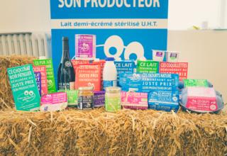 Différents produits alimentaires - produits laitiers, miel, oeufs, vin, compote de pommes, chocolat, etc. - de la marque "C'est qui le patron" disposés sur une botte de paille.