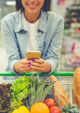 Photographie de consommateurs dans un super marché : au premier plan, une femme consulte son smartphone appuyée sur son caddie tandis qu'à l'arrière plan, un homme compare les étiquettes de deux produits.